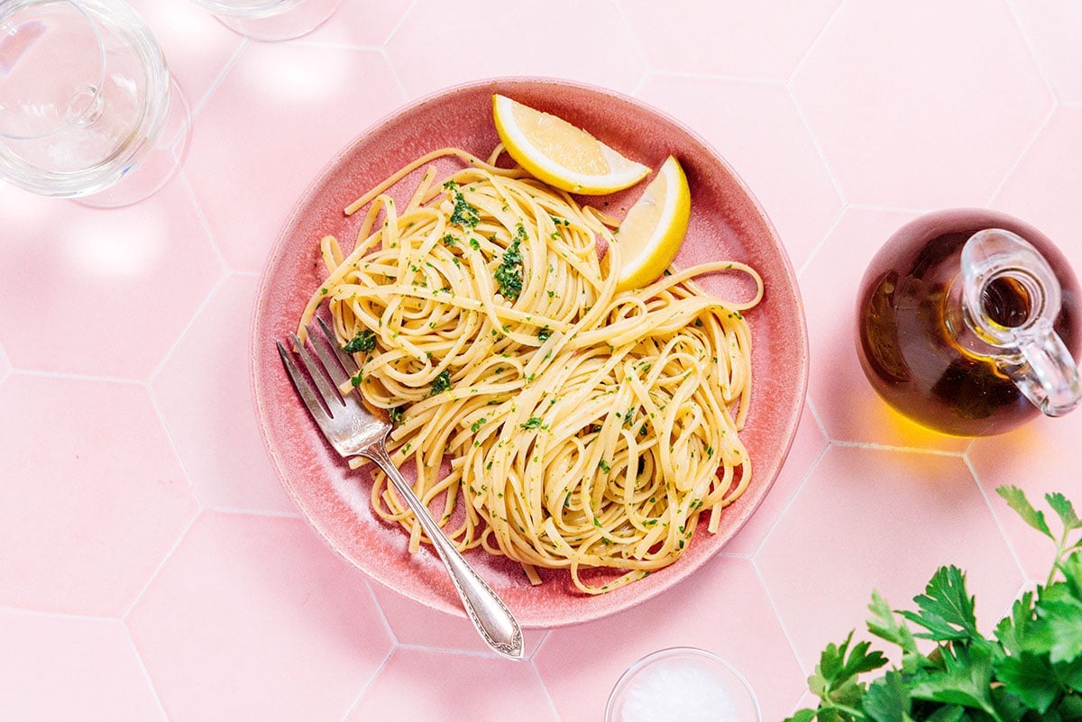 A pink bowl filled with lemon garlic pasta and two lemon wedges