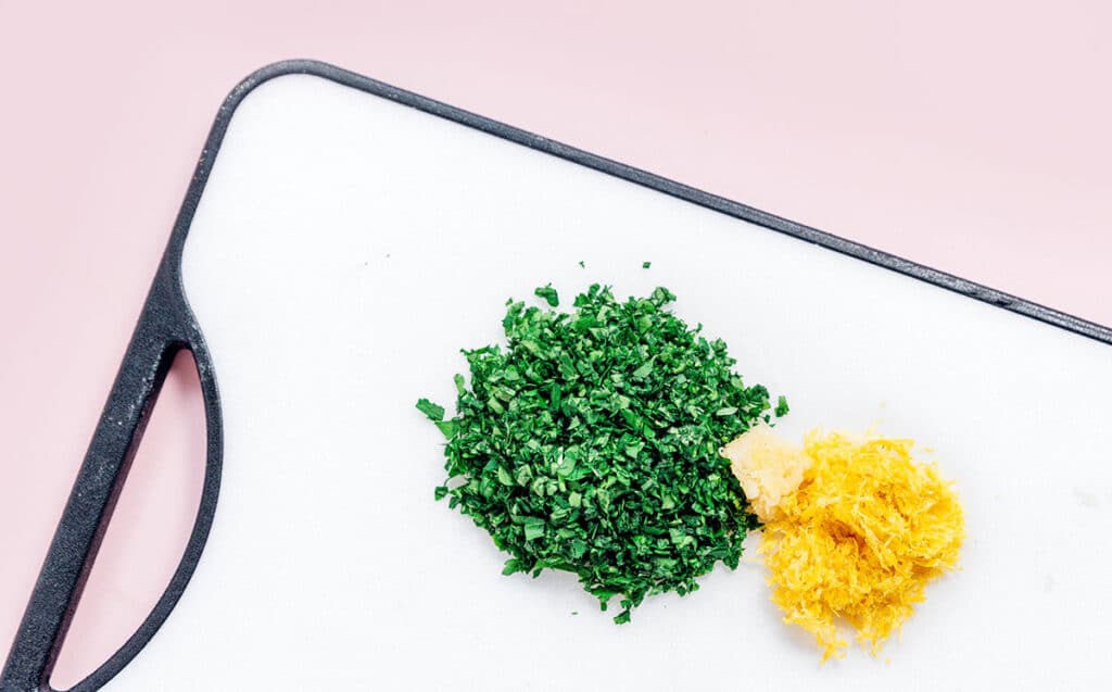 A cutting board topped with a small piles of chopped parsley, grated garlic, and lemon zest