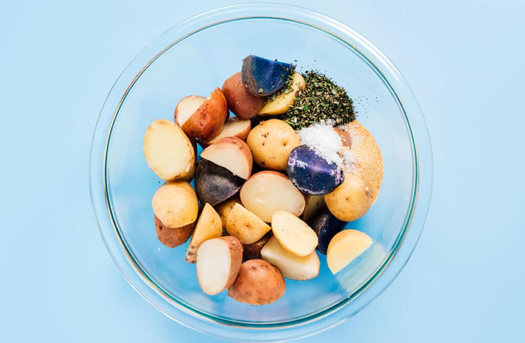 A clear glass bowl filled with potato halves, rosemary, garlic, salt, and oil