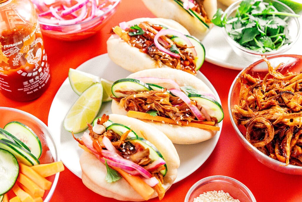 Assembled pulled mushroom bao buns arranged on white plates and surrounded with bowls of various ingredients like pickled red onion, cilantro, and cooked mushrooms