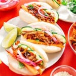 Plate of three vegan bao buns with spicy pulled mushrooms, cucumber, pickled onions, carrots, and sesame seeds. Lime wedges are on the side. Red background and additional dishes partially visible.