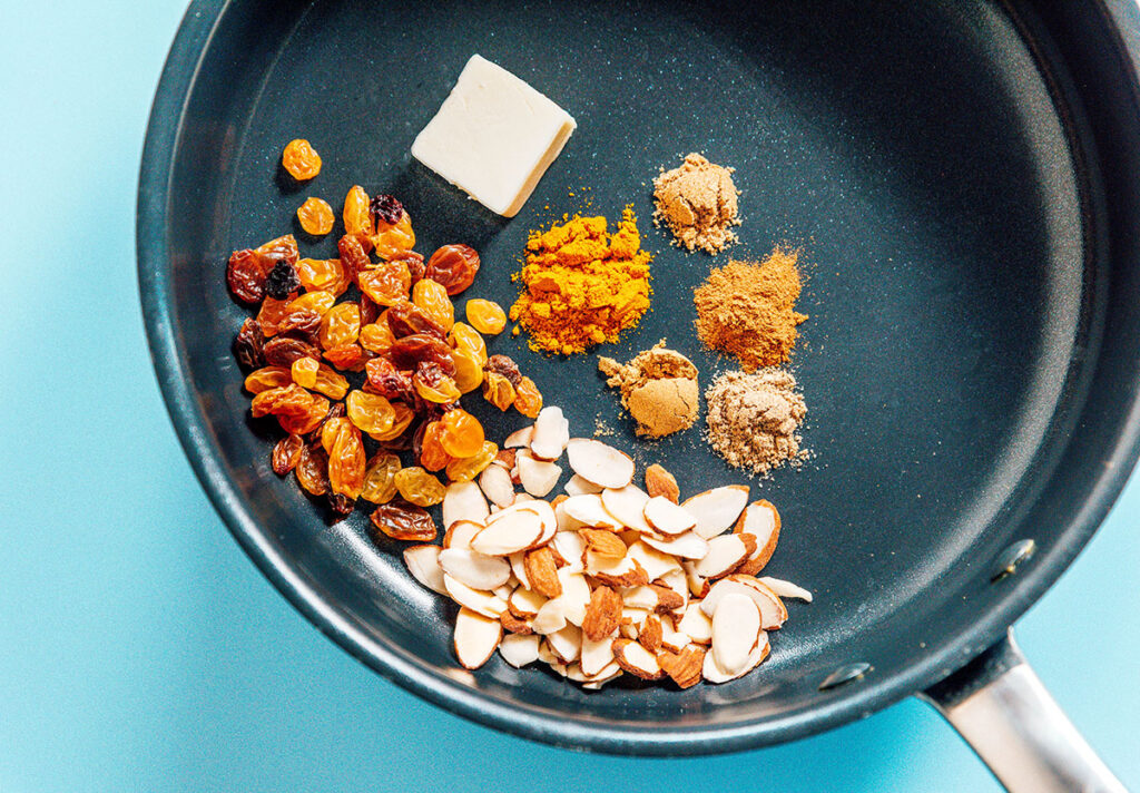 A skillet with golden raisins, sliced almonds, butter, and various spices