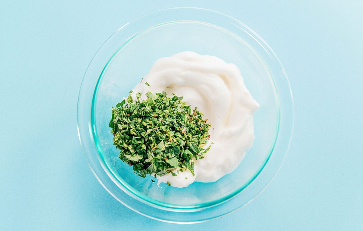 A clear glass bowl filled with biryani yogurt sauce ingredients: fresh mint, Greek yogurt, and salt