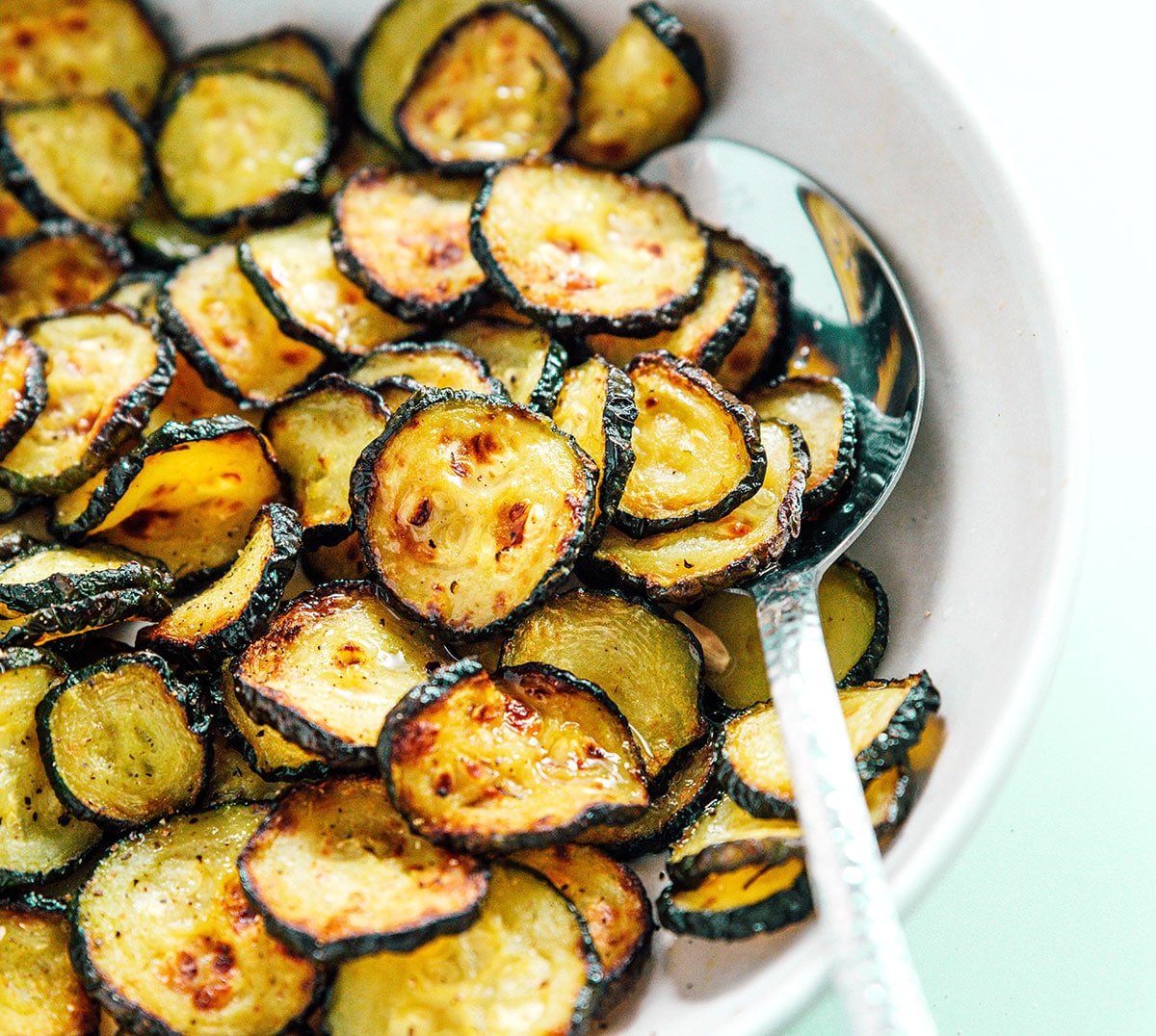 A silver spoon scooping air fried zucchini chips from a white bowl.