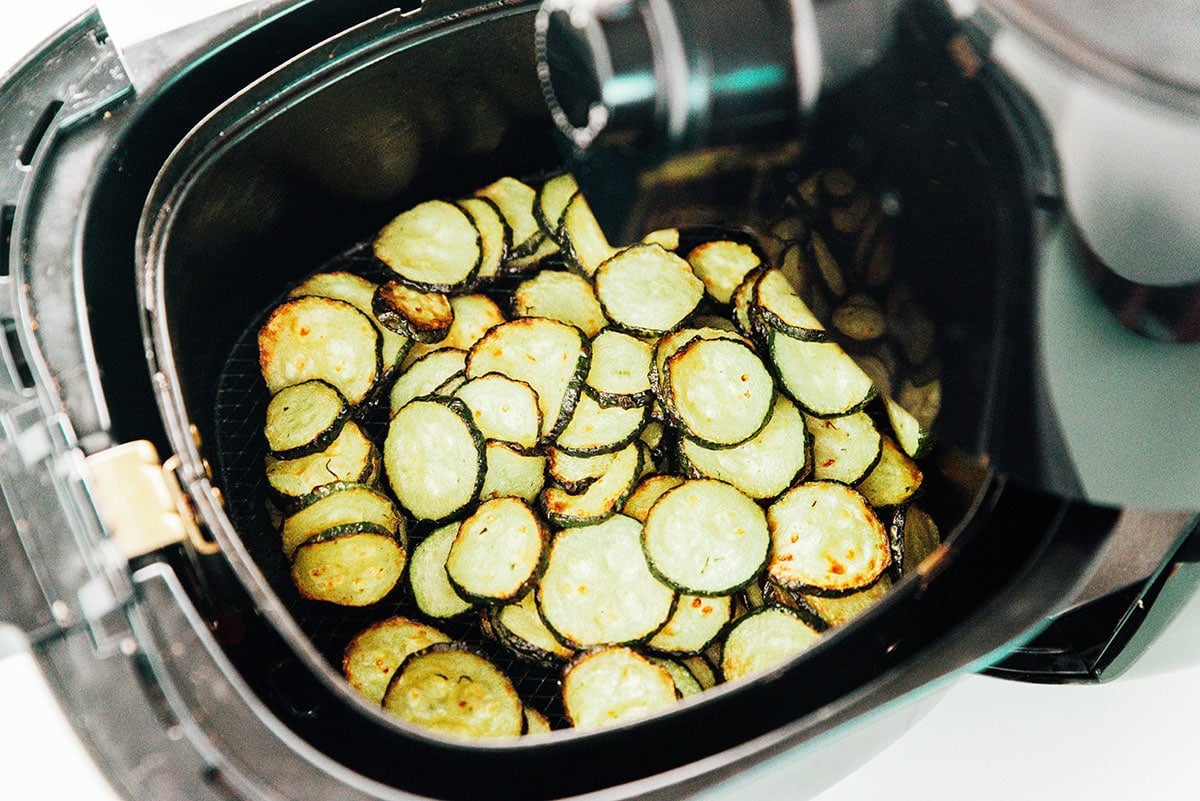 The basket being pulled from an air fryer, filled with zucchini rounds.