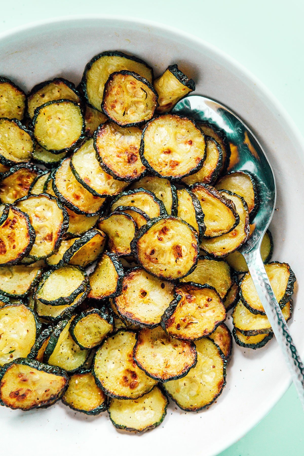 Air fryed zucchini chips with a silver spoon in a white bowl.