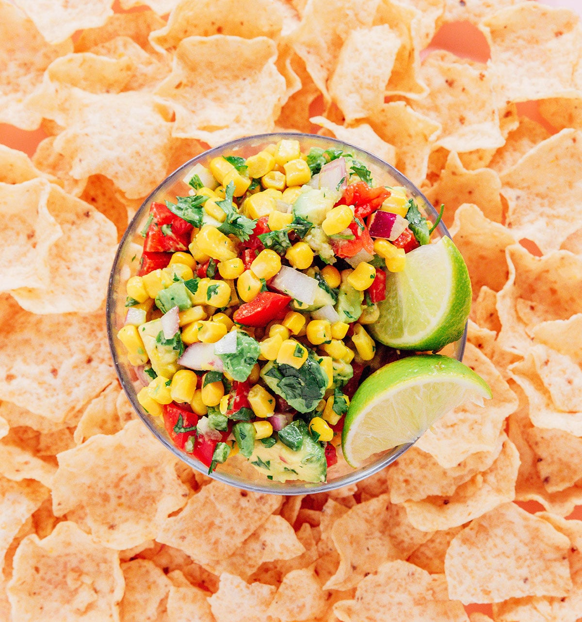 A small glass bowl of corn avocado salsa and lime wedges tucked into a bed of tortilla chips.