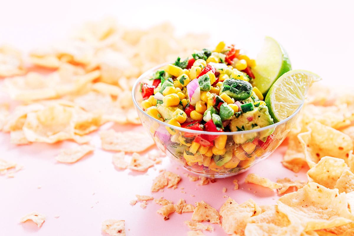 A small glass bowl of corn avocado salsa surrounded by chips on the table.