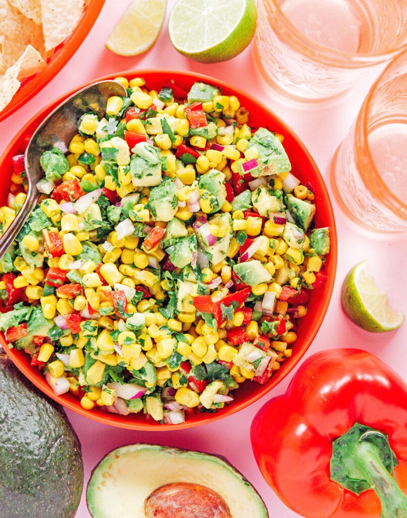 A red bowl filled with avocado corn salsa and a silver spoon.