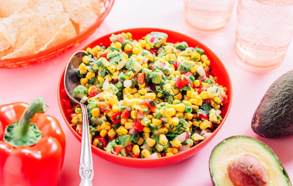 A red bowl filled with avocado corn salsa and a silver spoon.