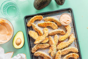Crispy avocado fries coated in breadcrumbs rest on a baking tray beside a zesty dipping sauce, with whole and halved avocados scattered across a vibrant green surface.