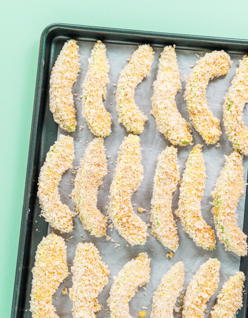 Breaded avocado slices lined up on a parchment paper lined baking sheet.