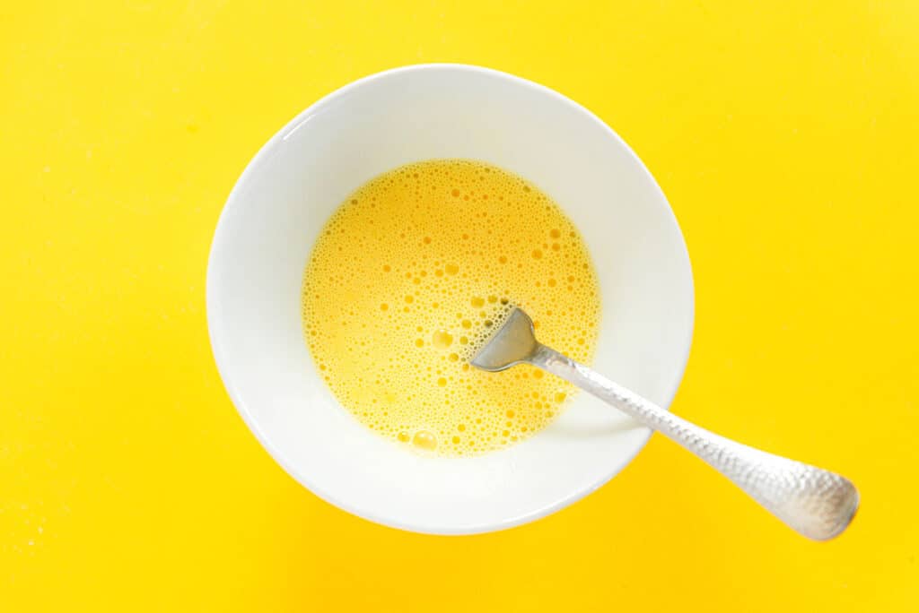 A fork in a white bowl of mixed eggs and milk.