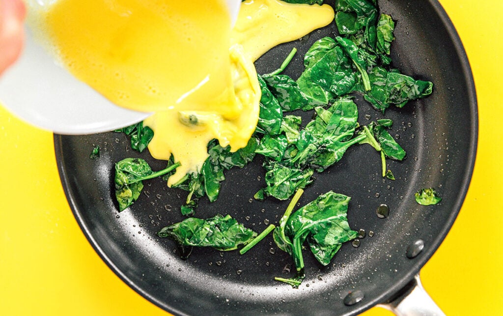 Whisked eggs being poured into a black skillet of sauteed spinach.
