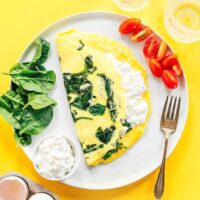 A cottage cheese omelette with fresh spinach is served alongside cherry tomatoes and a glass of water on a yellow background. A fork and a carton of eggs complete this vibrant, wholesome breakfast scene.