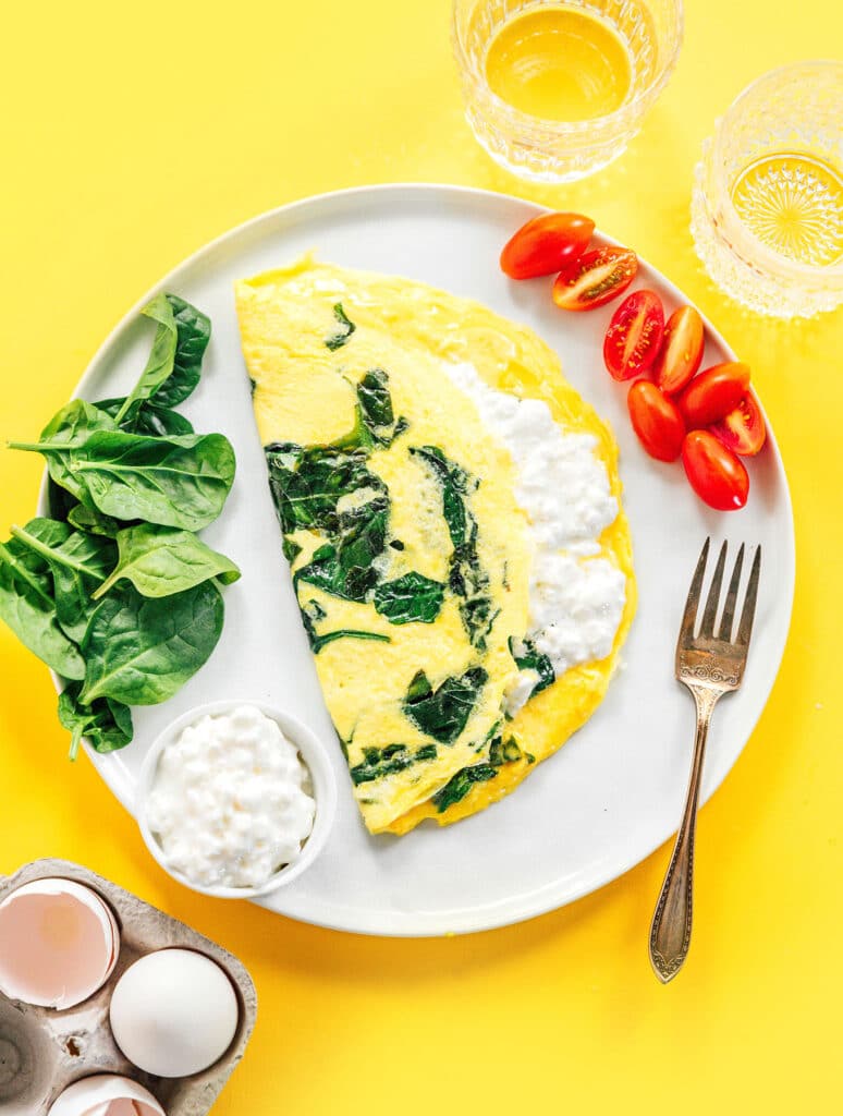 A large white plate with a spinach and cottage cheese omelette next to spinach and cherry tomatoes.