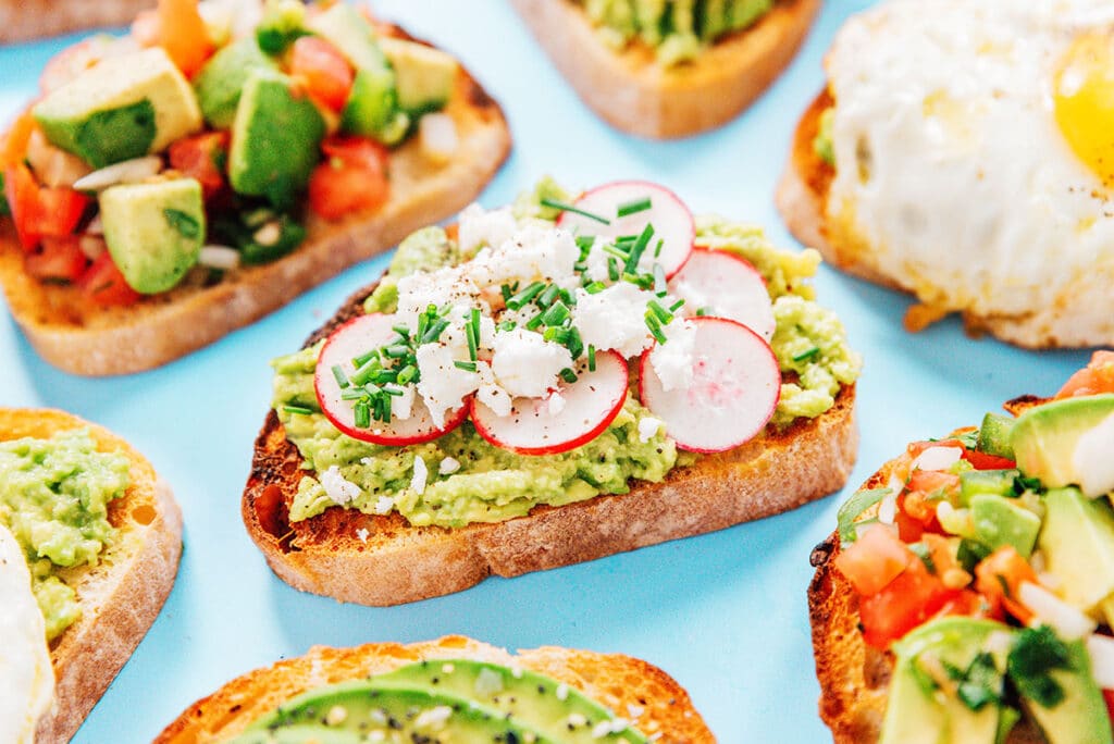 A slice of goat cheese and chive avocado toast surrounded by other avocado toast variations on a blue background.