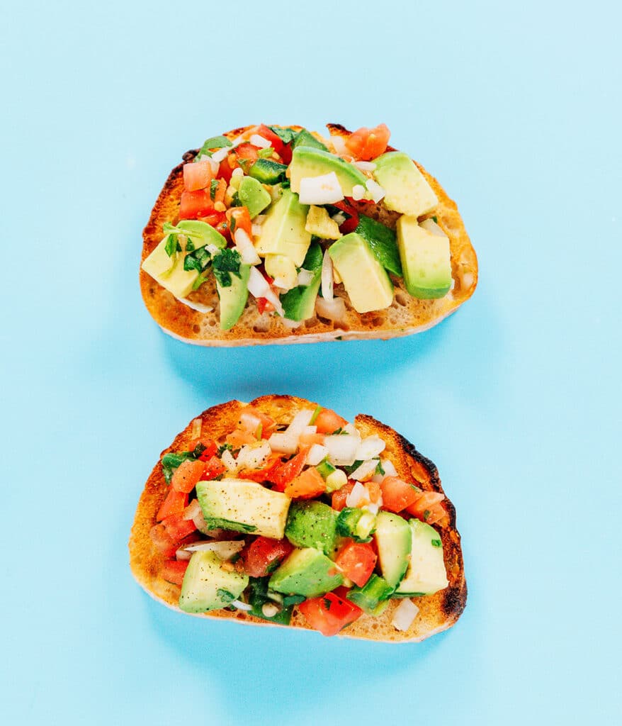 The pico de gallo toast with diced avocado toast and pico de gallo on two slices of bread  on a blue backdrop.