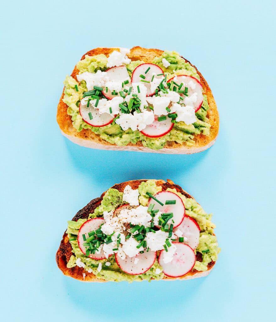 Two pieces of goat chees and chive avocado toast with mashed avocado, radishes, goat cheese and chives on a blue back ground.