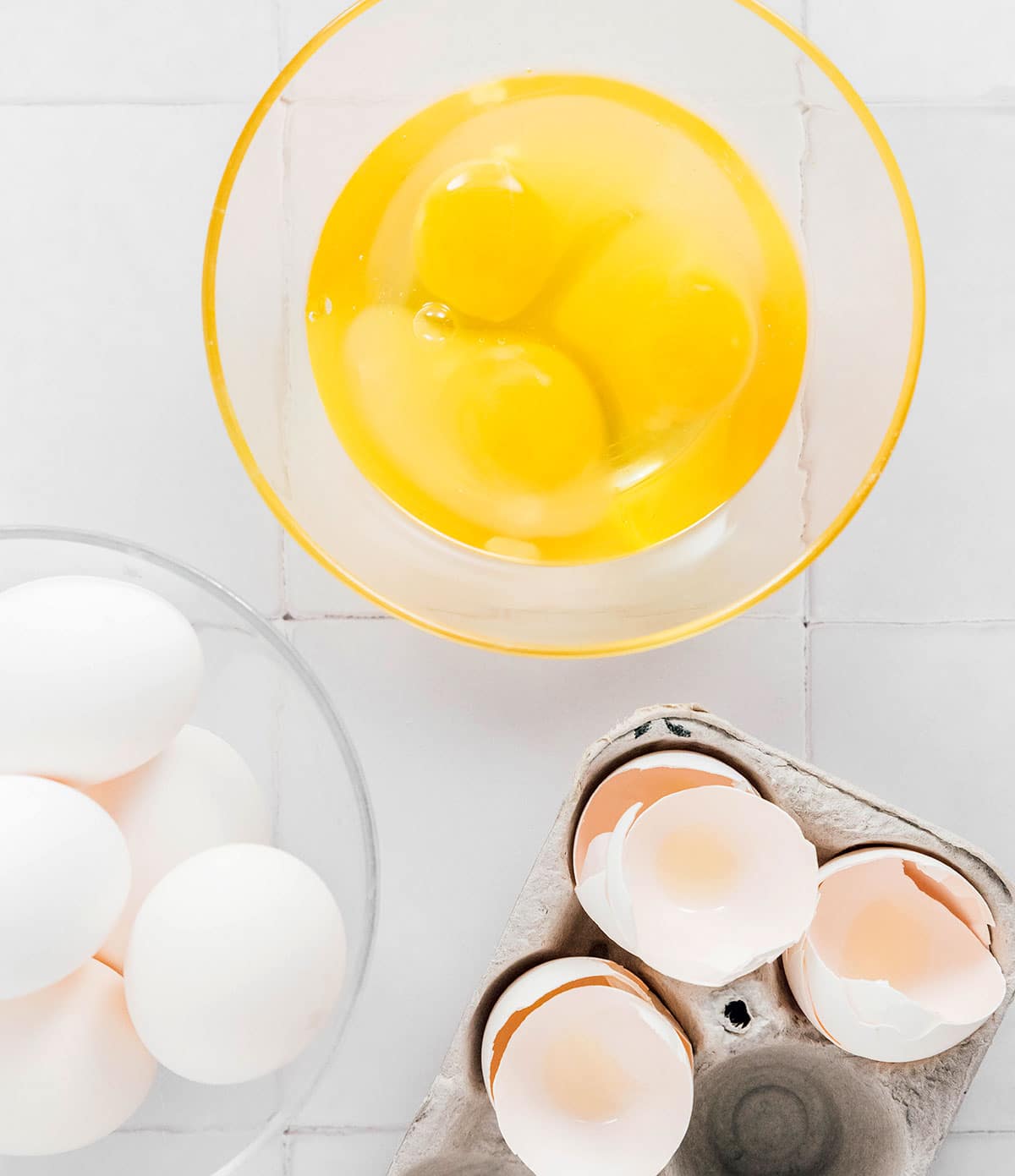 A bowl of cracked eggs next to a bowl of whole eggs, sitting by a carton of egg shells.