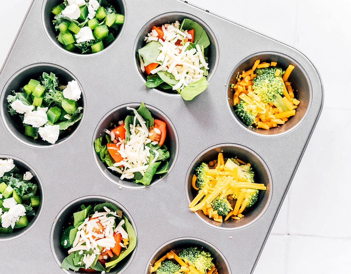 Chopped veggies and shredded cheese filling a greased metal muffin tin.