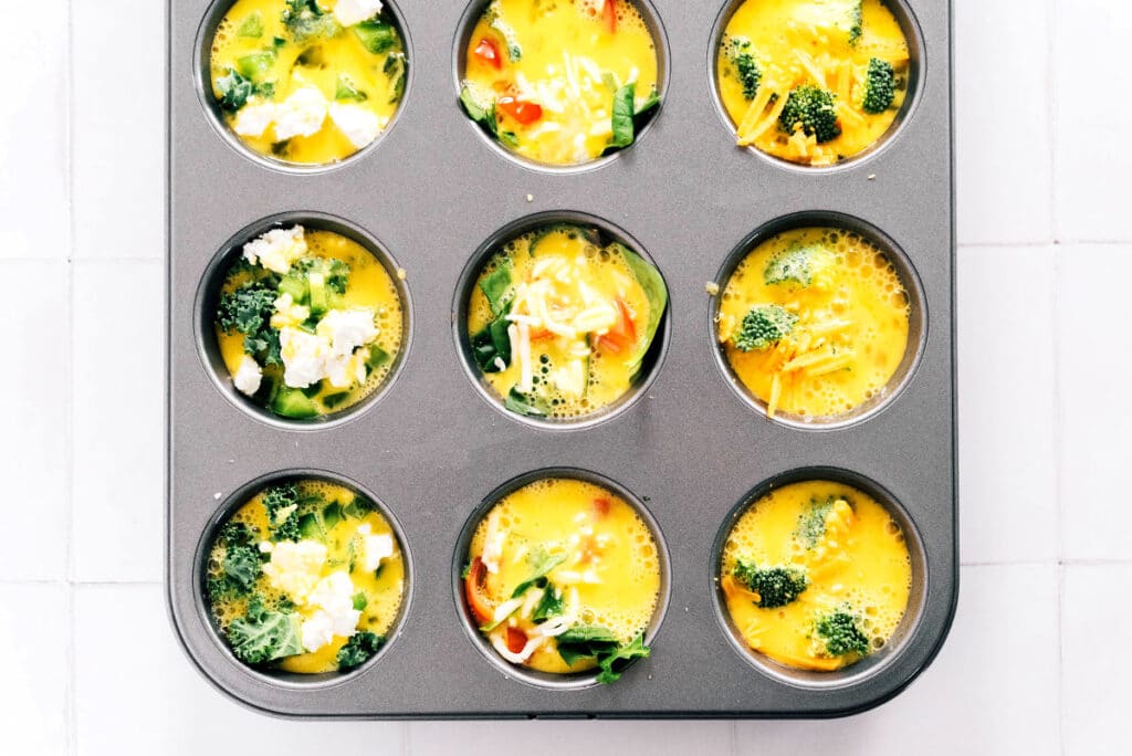 Beaten eggs poured over veggies and cheese in a metal muffin tin.