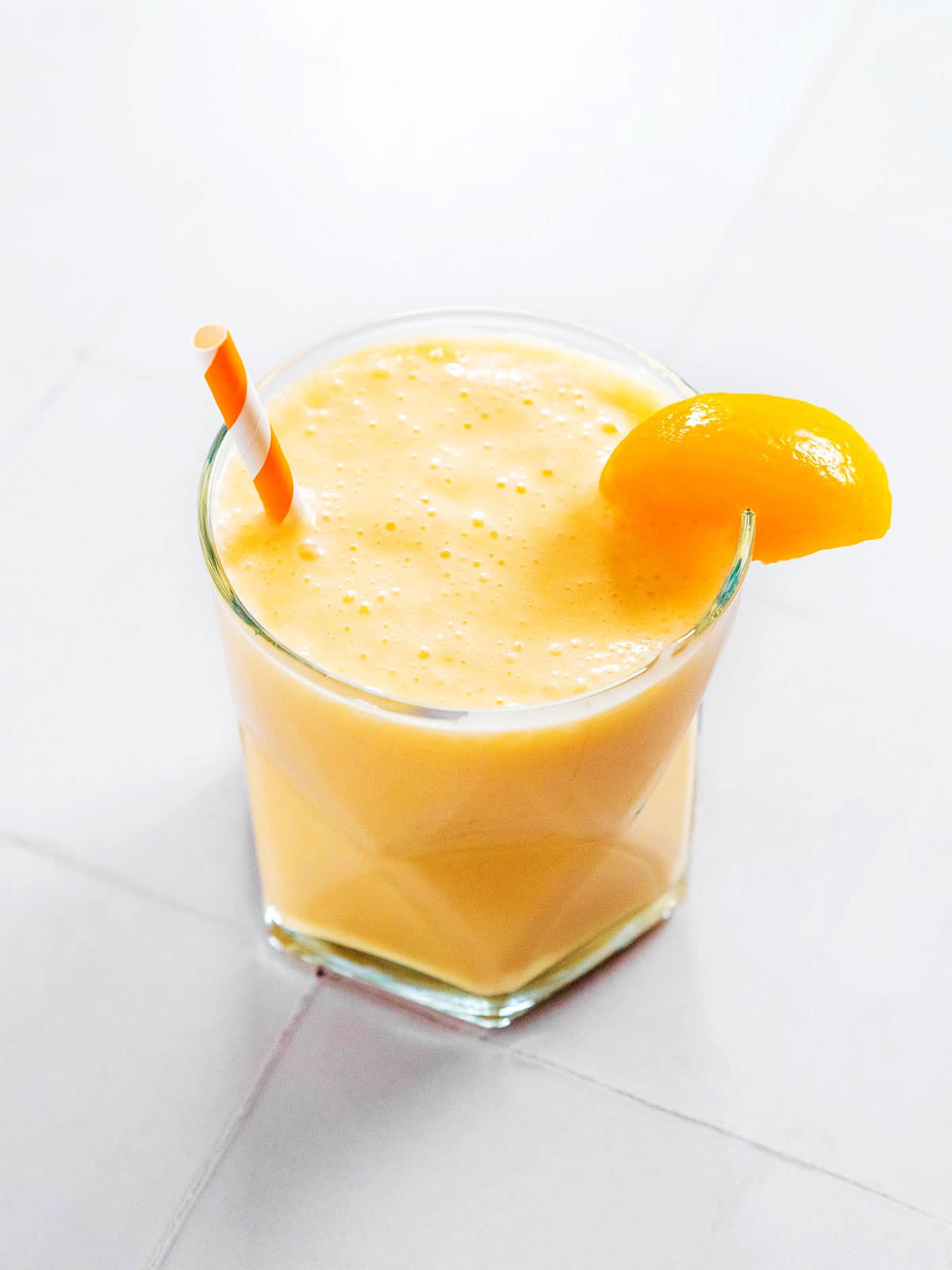 A small glass of peach kefir with a straw and a peach slice on the rim.