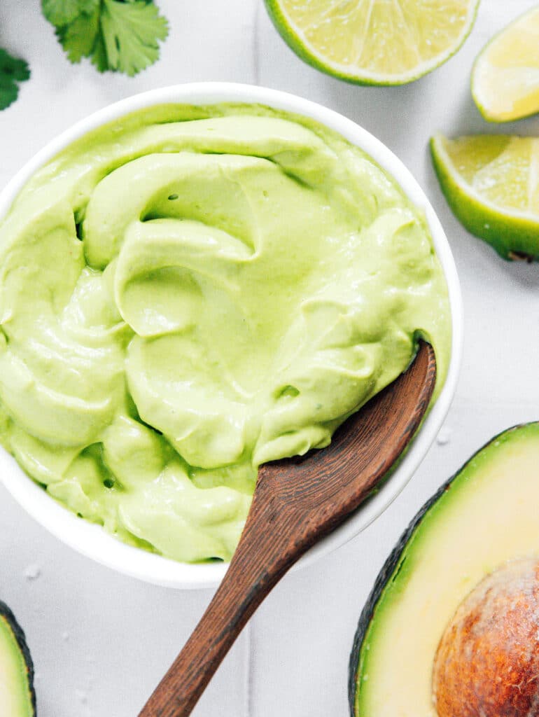 Small bowl of avocado crema and a wooden spoon with halved avocados next to it.