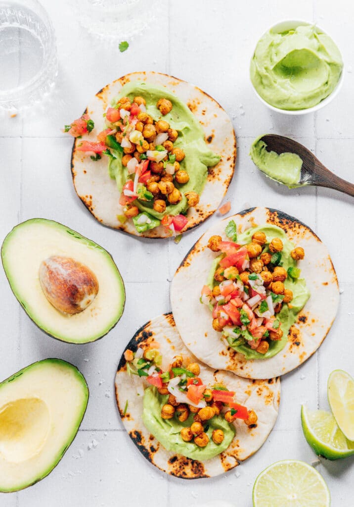 Chickpea tacos with avocado crema on a tile surface with avocados and limes next to them.
