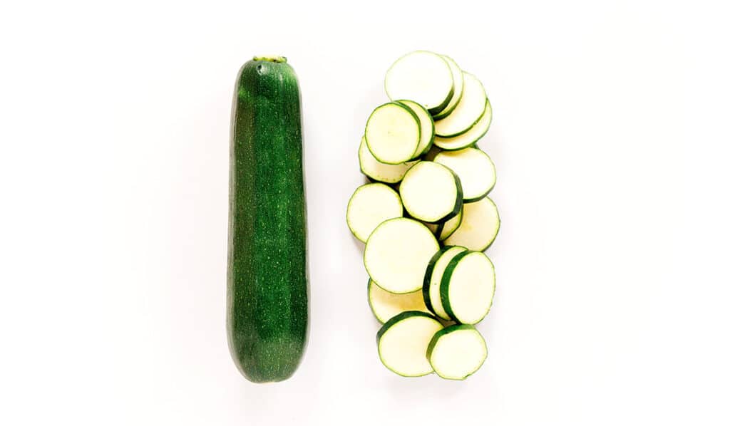 A single zucchini next to a pile of sliced zucchini rounds.