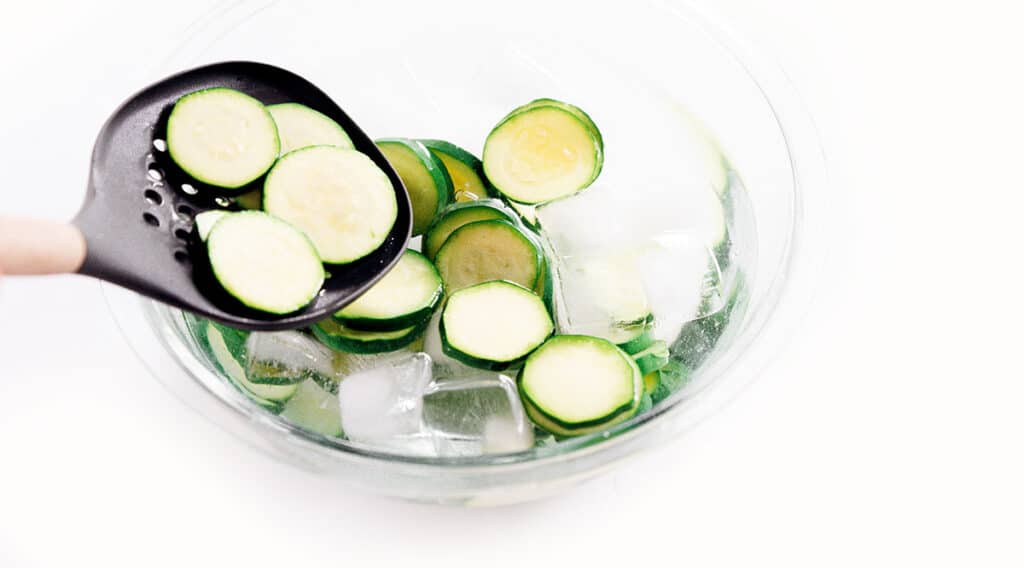 A slotted spoon placing blanched zucchini rounds into a bowl of ice water.