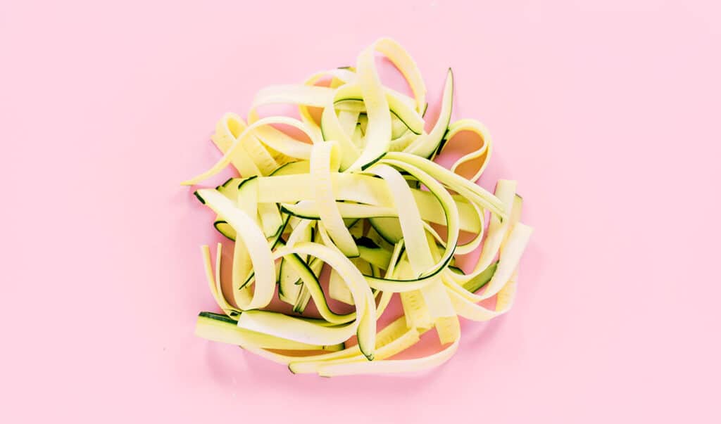 A pile of fettuccine zucchini noodles n a pink background.