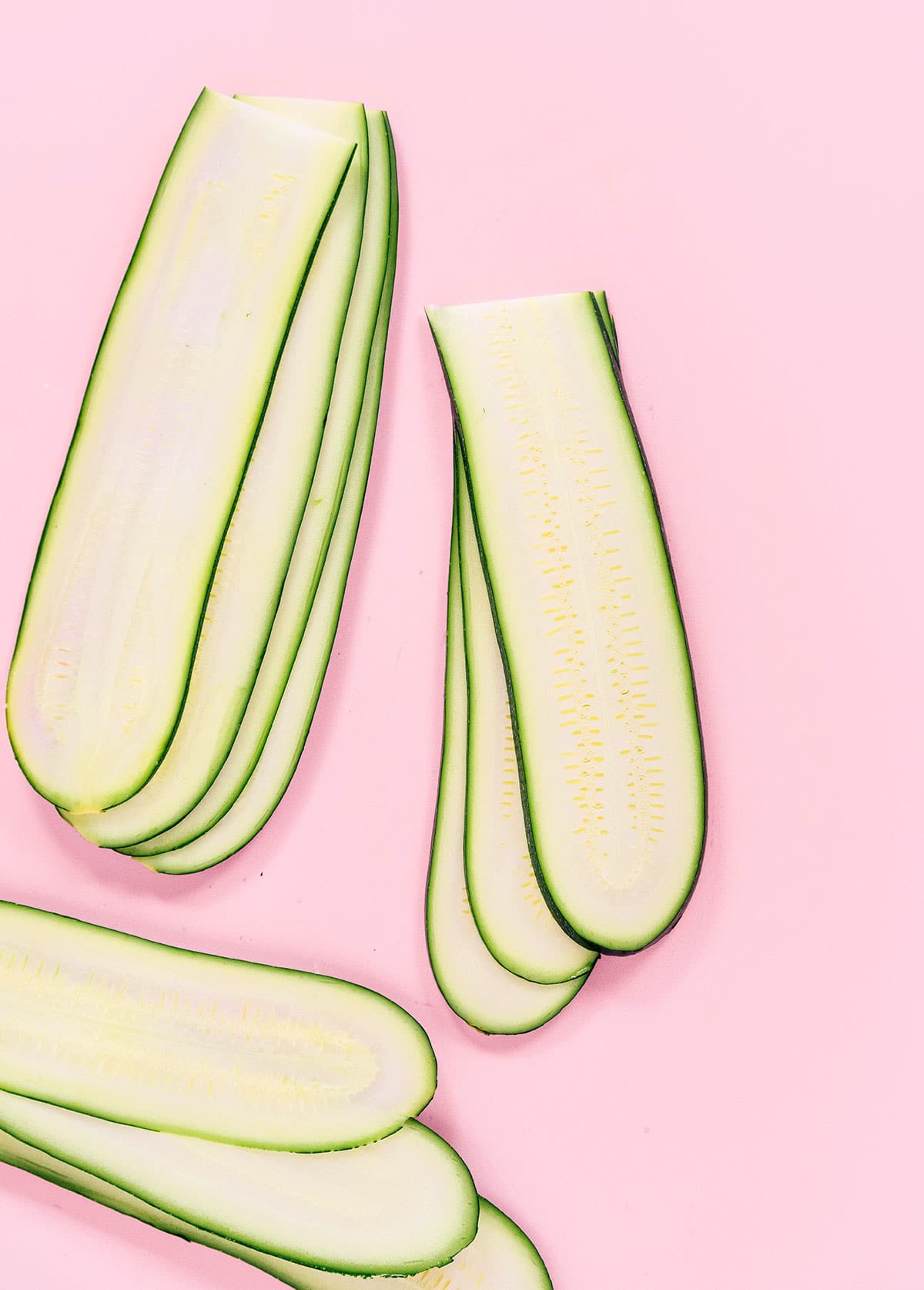 Multiple thin, lengthwise slices of zucchini laid flat on a pink back ground.