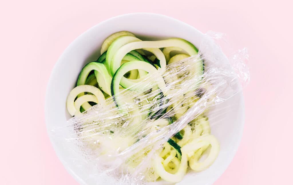 A white bowl on a pink background filled with spaghetti zoodles and partially covered in plastic wrap.  As if the bowl is being covered by plastic wrap.