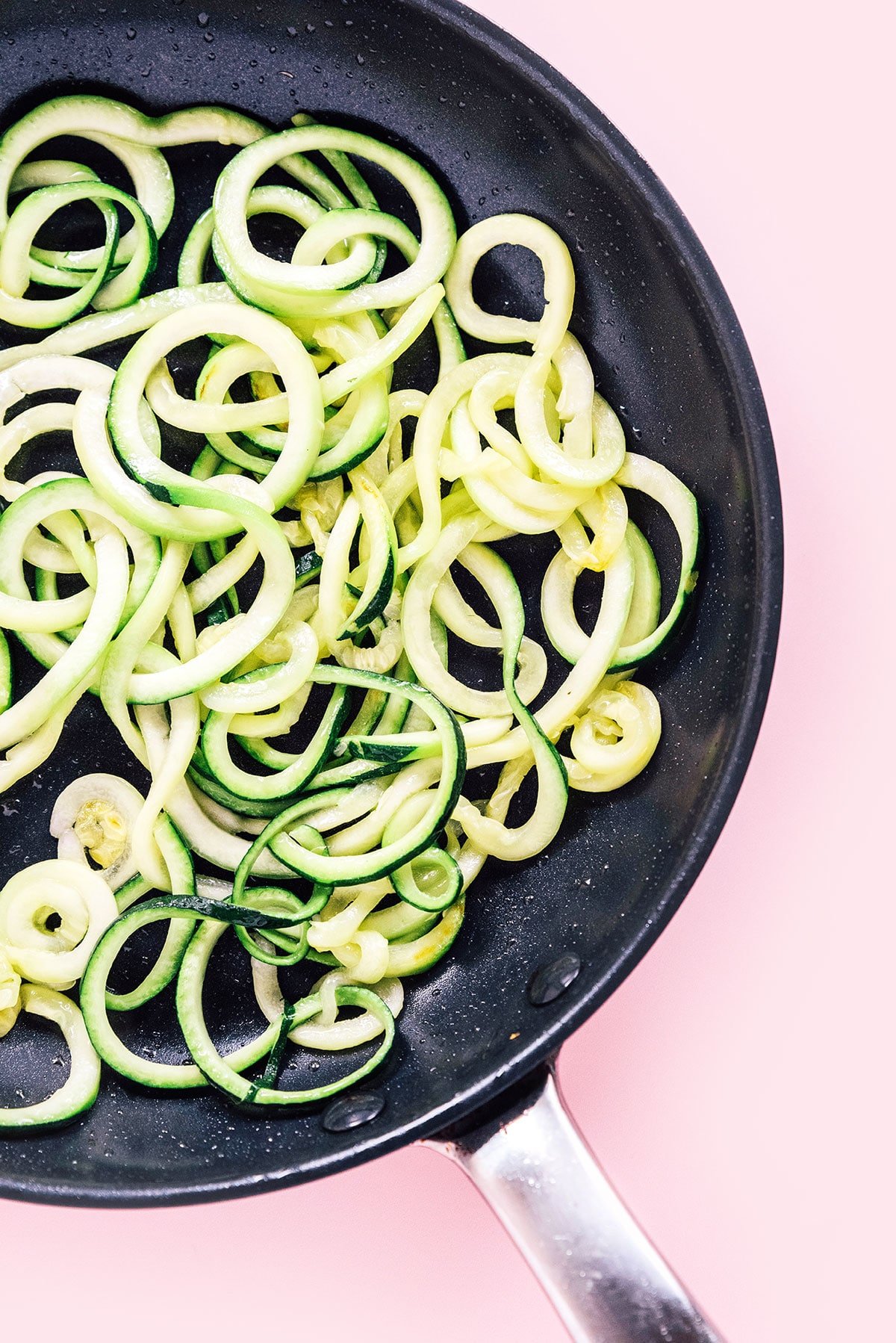 A black pan with spaghetti zoodles in in on a pink background.