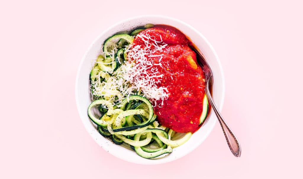 Spaghetti zoodles, red sauce, parmesan, and a fork in a white bowl on a pink background.