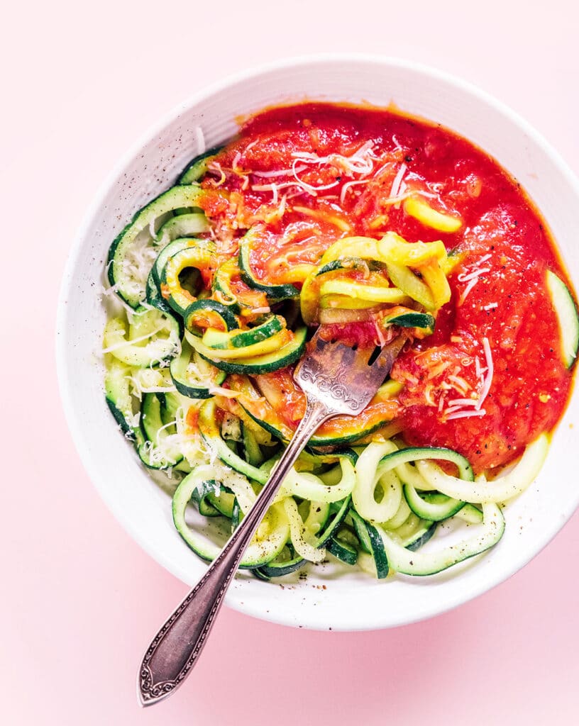 Spaghetti zucchini noodles with red sauce, parmesan cheese, and a fork in a white bowl.