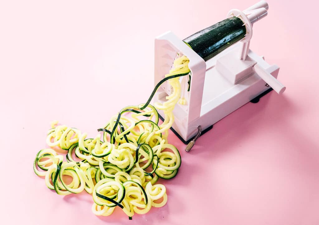 A spiralizer with half of a zucchini it in and the other half pushed through the spiralizer and turned into spaghetti zoodles in a pile on a pink background.