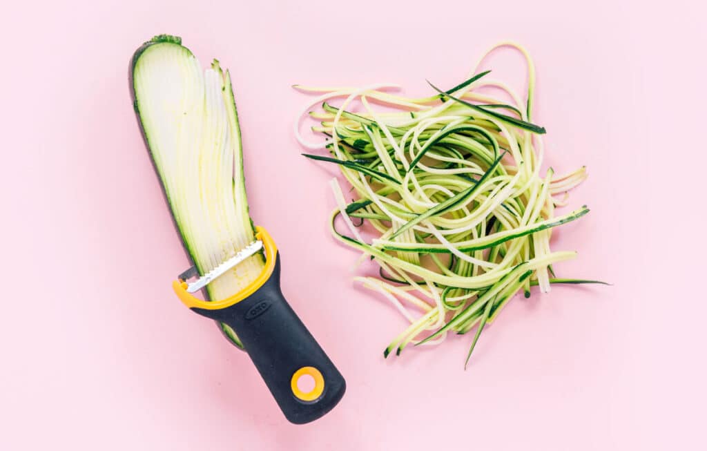 A pile of angle hair zucchini noodles next to half of a zucchini that is being sliced by a black and yellow julienne slicer.