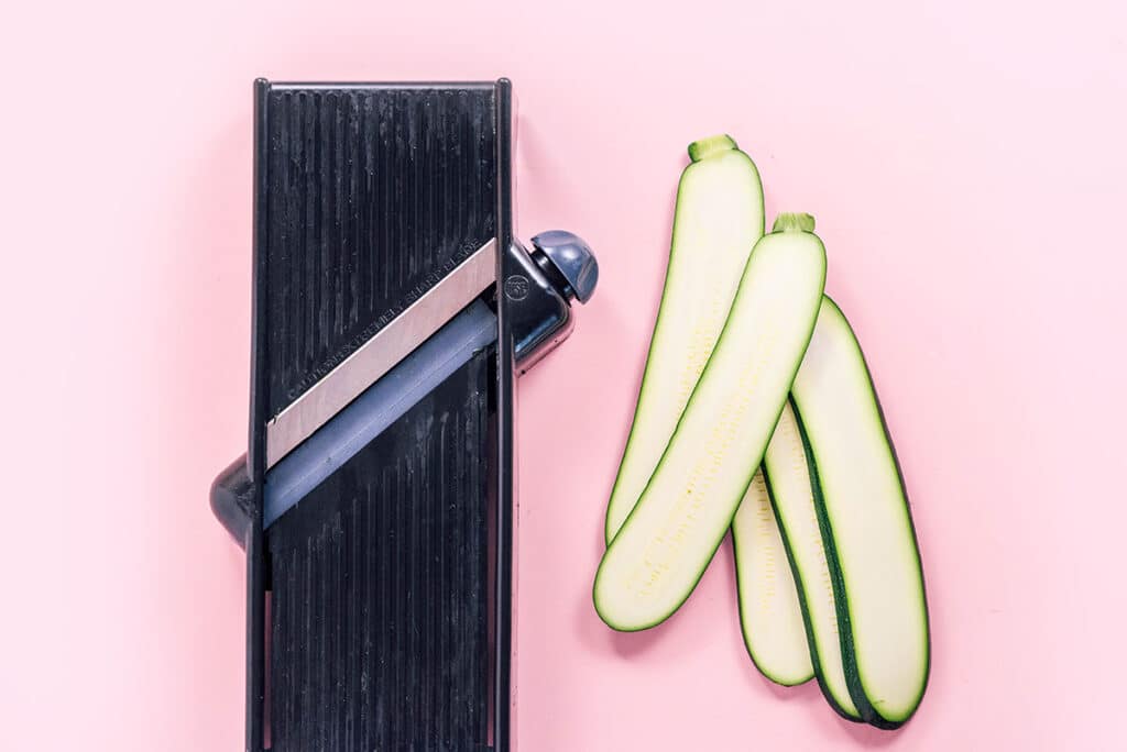 A mandolin slicer next to 5 lengthwise slices of zucchini on a pink background.