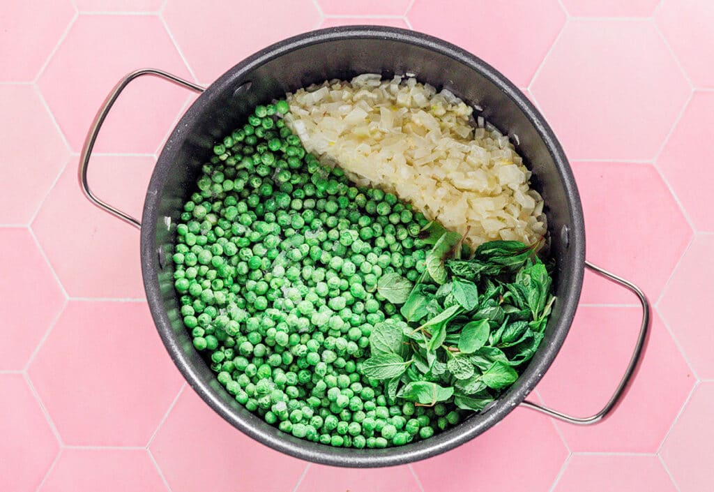 A cooking pot filled with sautéed white onion, peas, and mint leaves