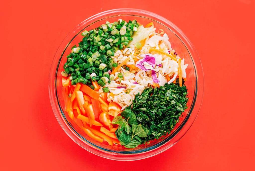 A clear glass bowl filled with Thai cabbage salad ingredients including coleslaw, green onions, red bell pepper, cilantro, and mint