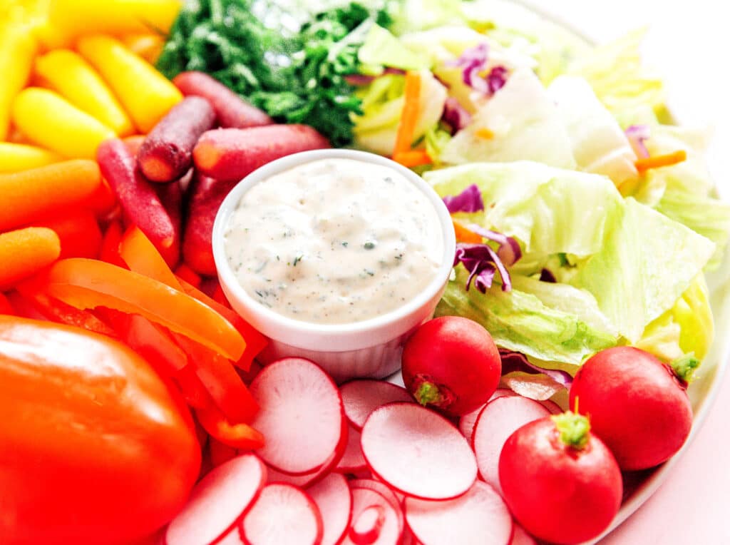 A platter filled with a bowl of vegan ranch dressing surrounded by veggies like peppers, carrots, radishes, lettuce, and more