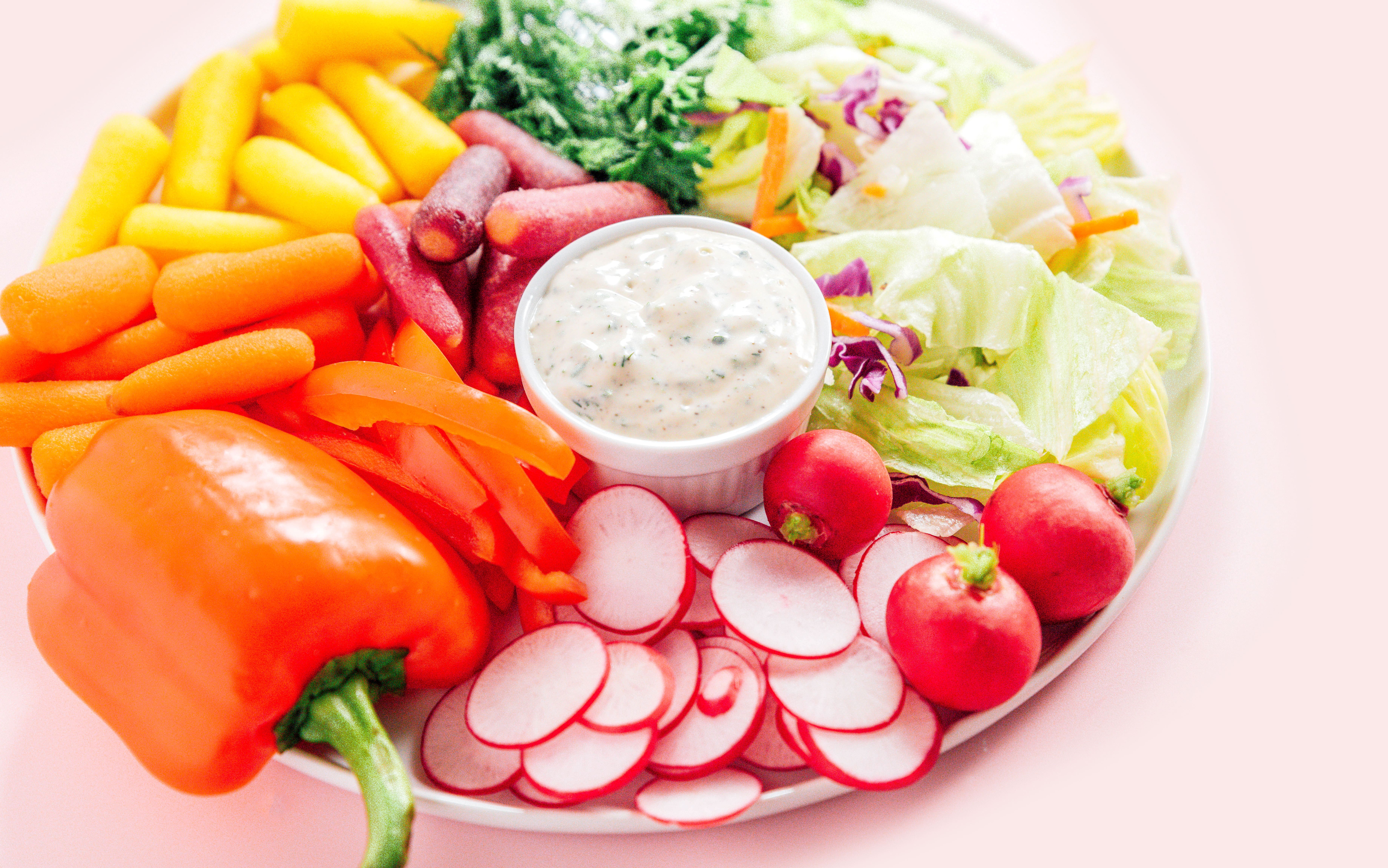 A platter filled with a bowl of vegan ranch dressing surrounded by veggies like peppers, carrots, radishes, lettuce, and more