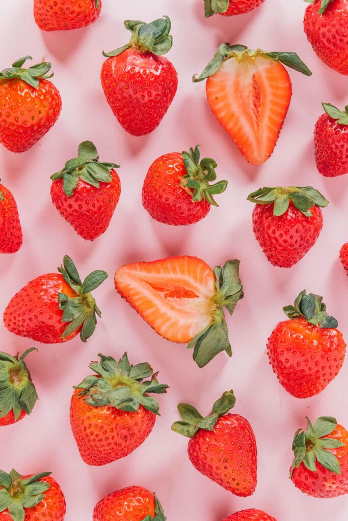 Whole strawberries with a few sliced in half spread out on a pink surface.