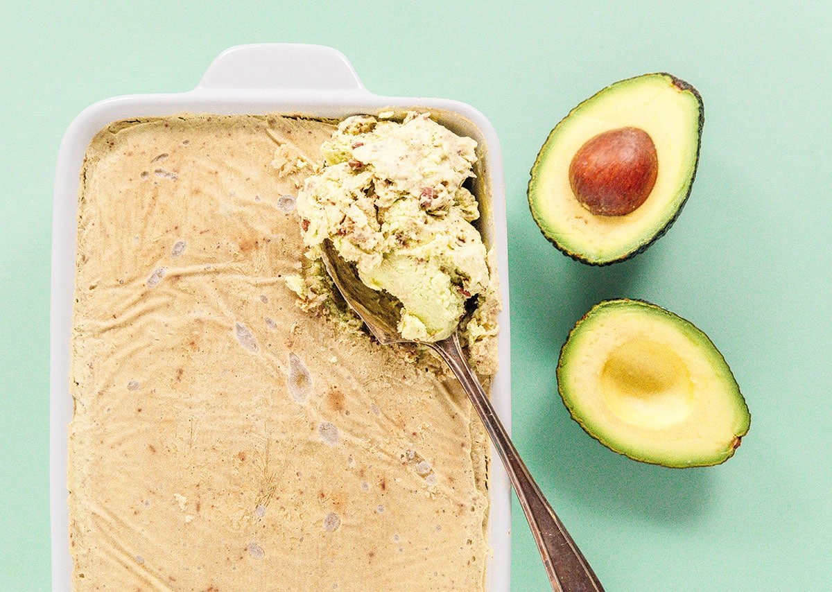 Ice cream in the baking dish with a scoop being taken out and a sliced open avocado sitting next to it.