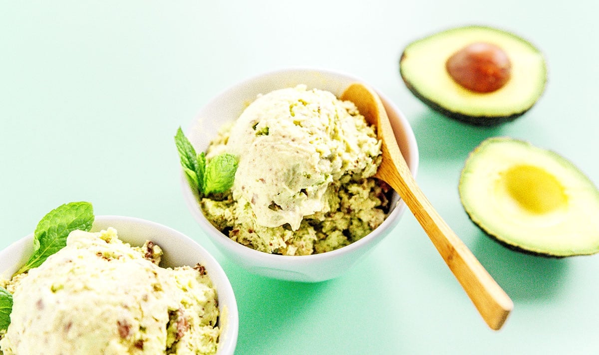Two bowls of mint chocolate avocado ice cream with a wooden spoon in one of the bowl and an open avocado sitting next to them.