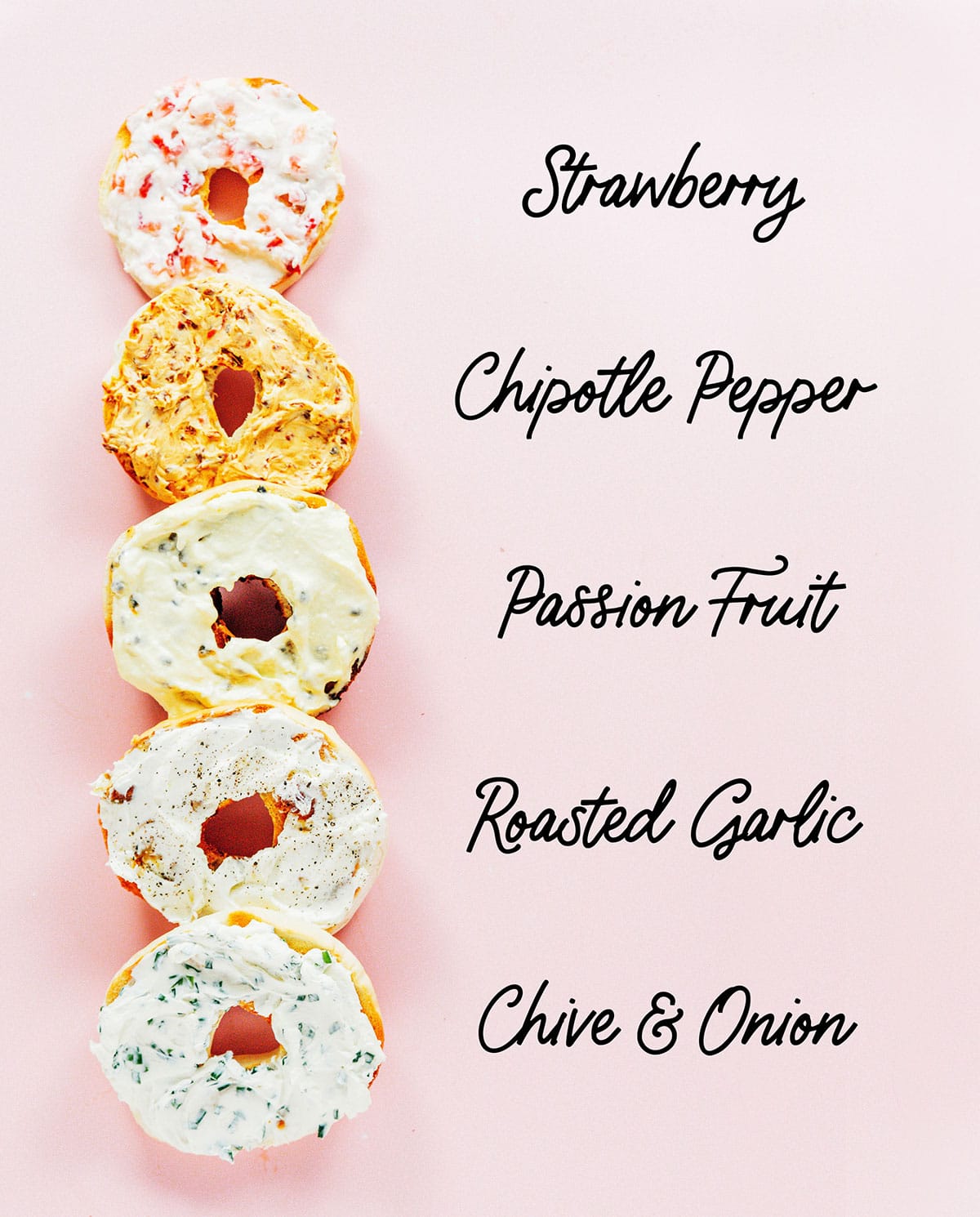Bagel halves spread with various flavors of cream cheese on a pink surface.