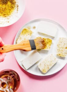 A hand uses a brush to spread oil and seasoning onto rectangular blocks of grilled halloumi arranged on a white plate.