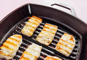 Six pieces of white fish and grilled halloumi are being cooked on a ridged cast iron grill pan, with visible grill marks and light seasoning.